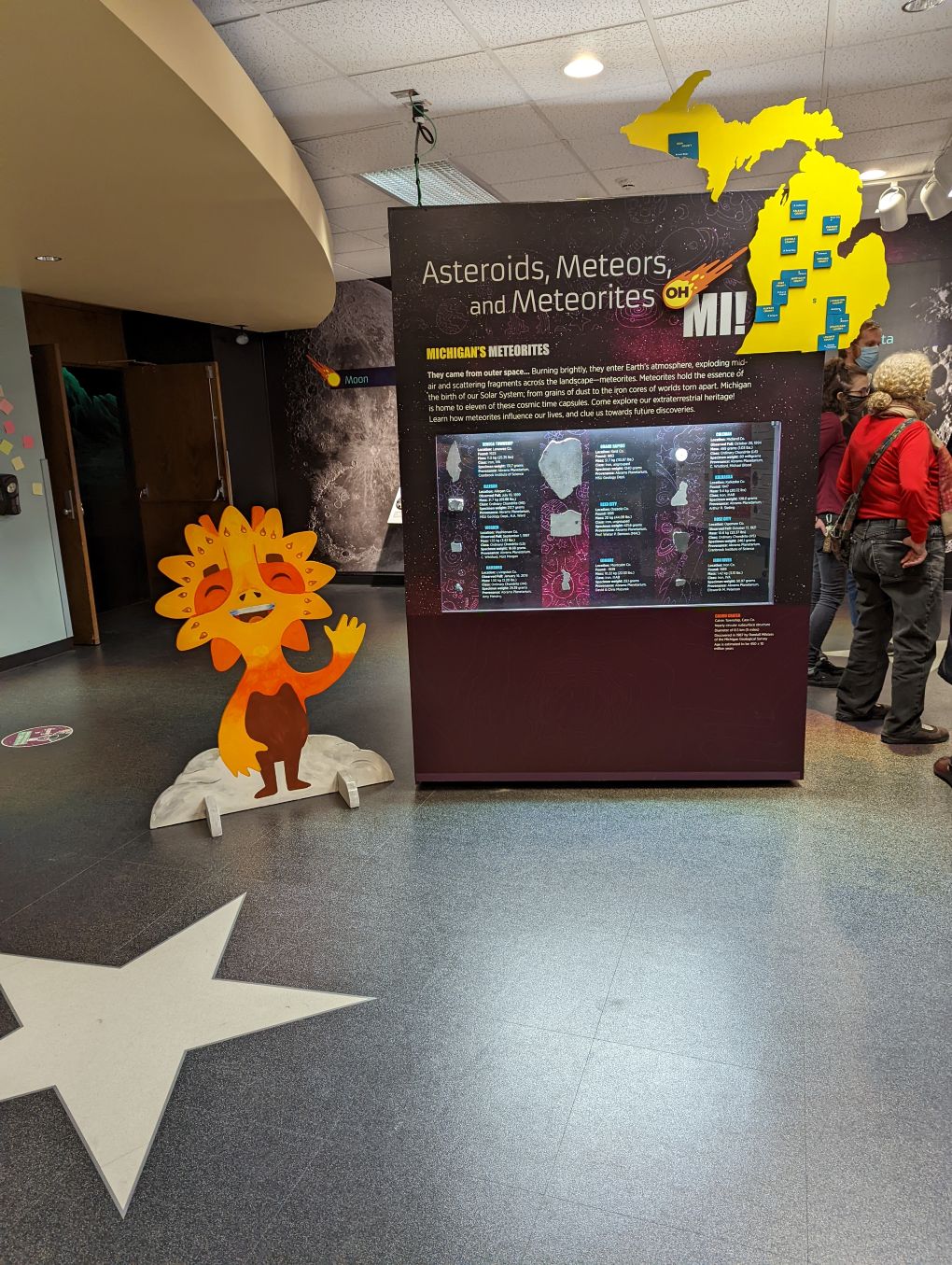 Image of meteorite exhibit with large cabinet with a yellow cut-out of Michigan on top and several rocks mounted in the middle behind glass. People browse to the right and there is a cutout of a yellow and orange alien creature to the left.