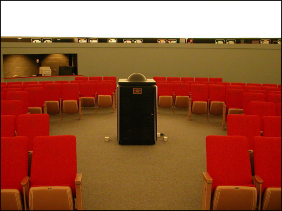 Picture of theater with bright red seats and a boxy projector in teh center with a large lens