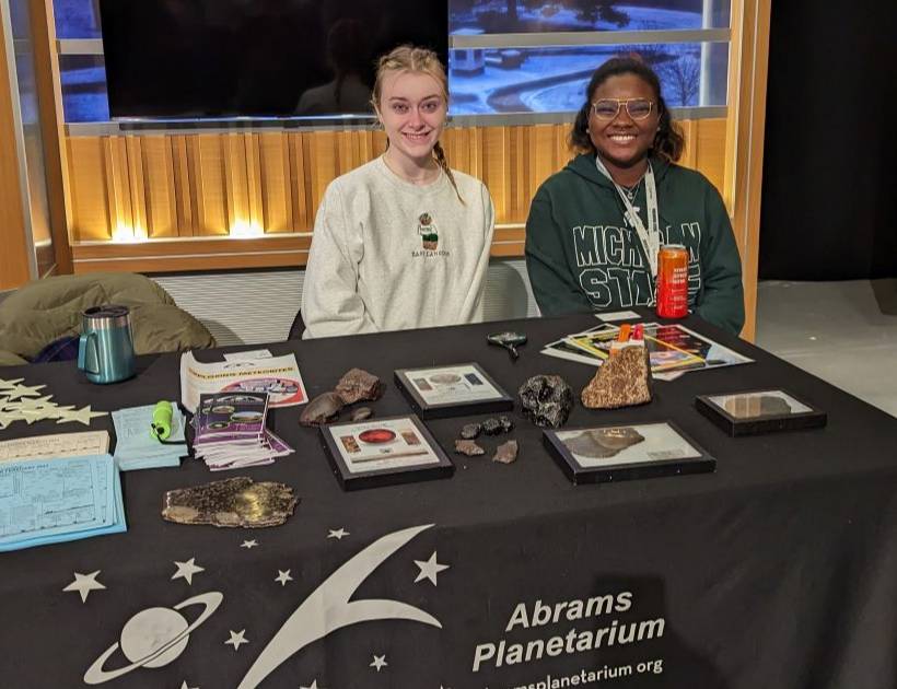 A young black woman and young white woman sit behind a table smiling with meteorites, flyers, and stickers at an event table..