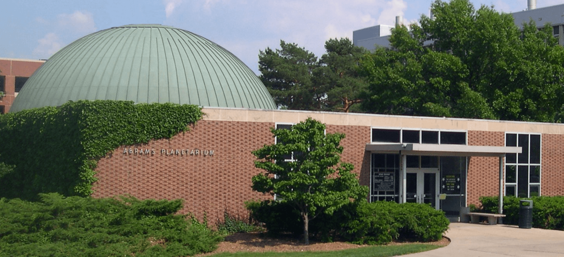 Color image of planetarium building, a boxy red brick building with copper dome in a teal patina surrounded by bushes and trees.