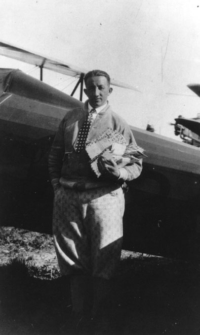 Black and white photo of a young Ted Abrams in front of a plane in slacks and a jacket holding an item.